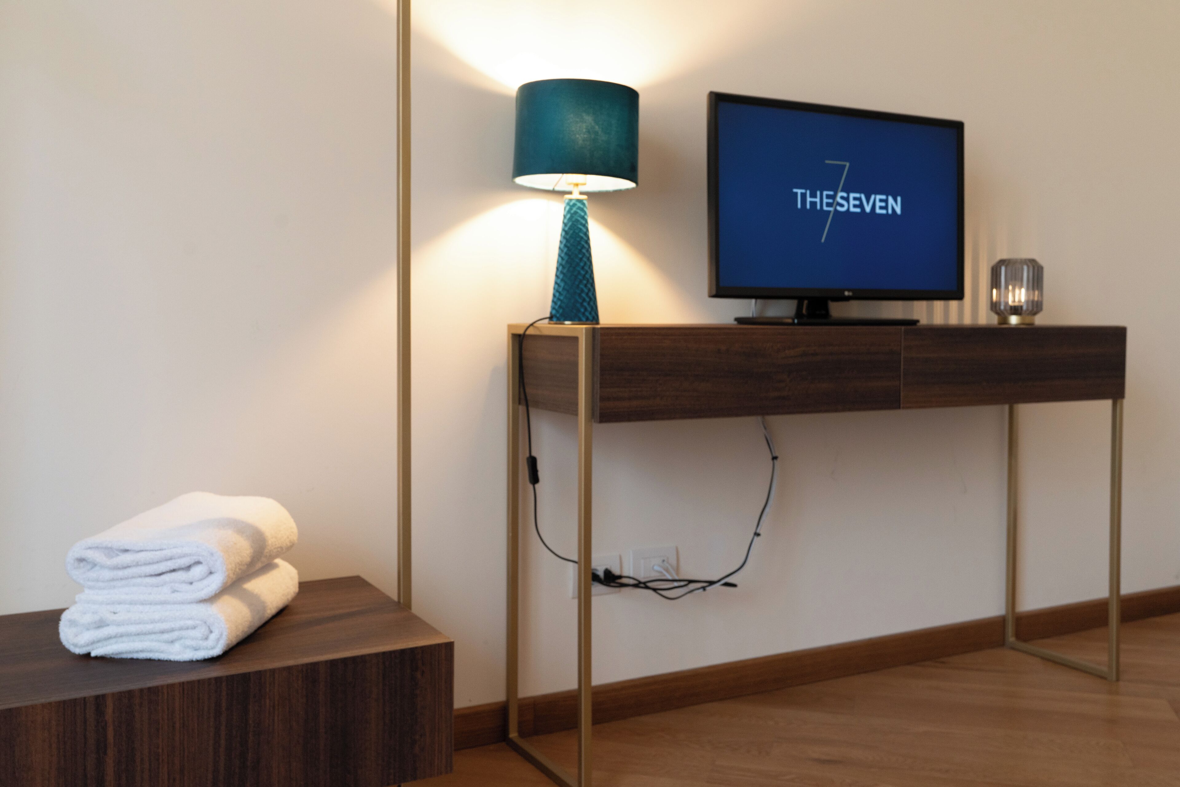 A modern hotel room with a wooden console table, a television displaying 'THESEVEN', and a lamp.