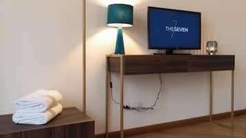 A modern hotel room with a wooden console table, a television displaying 'THESEVEN', and a lamp.