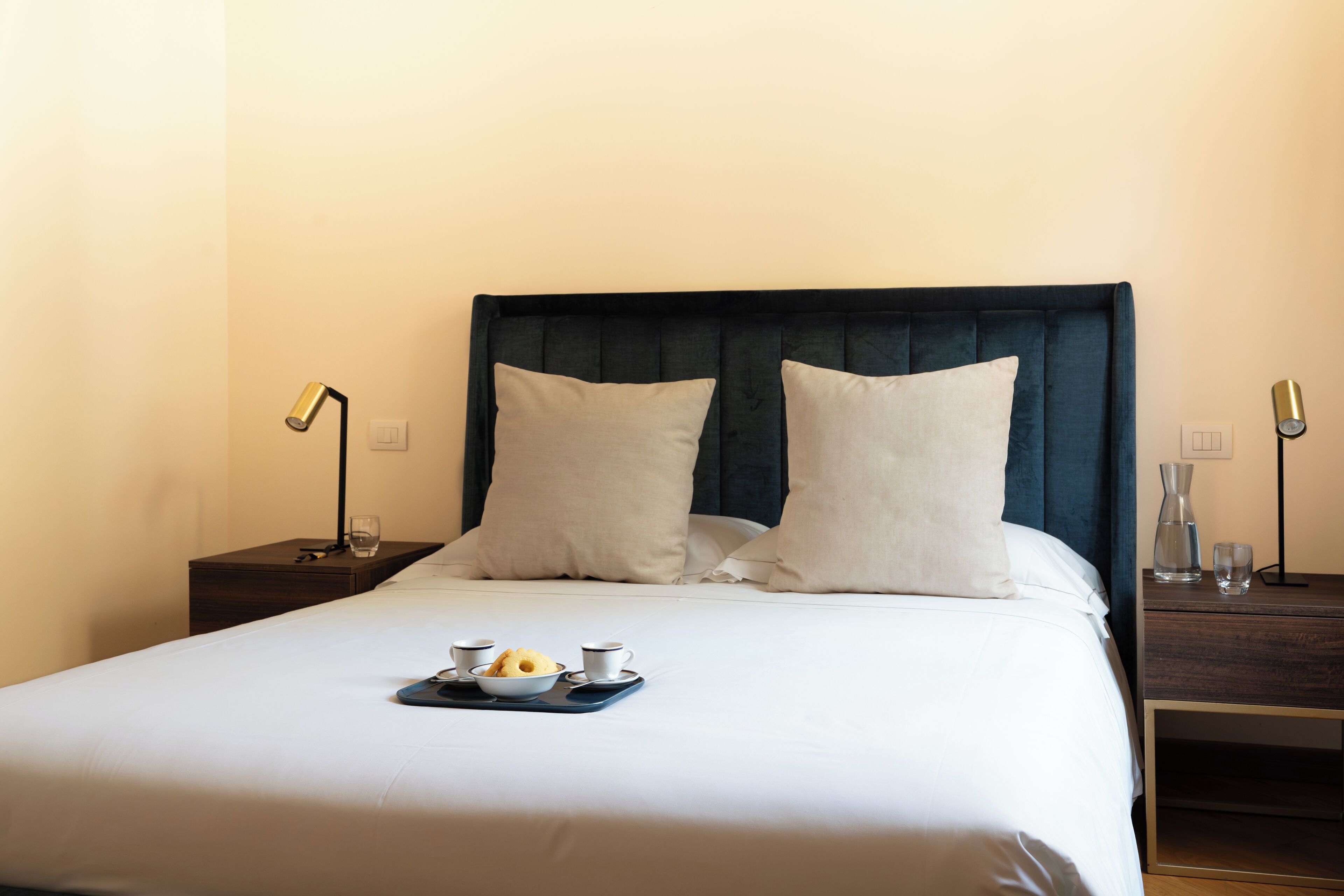 A neatly made bed with a tray of breakfast items, two bedside lamps, and a dark headboard in a hotel room.