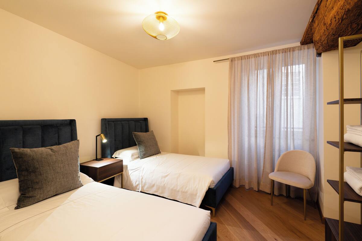 A hotel room with two beds, a chair, a desk with a lamp, and a wardrobe.