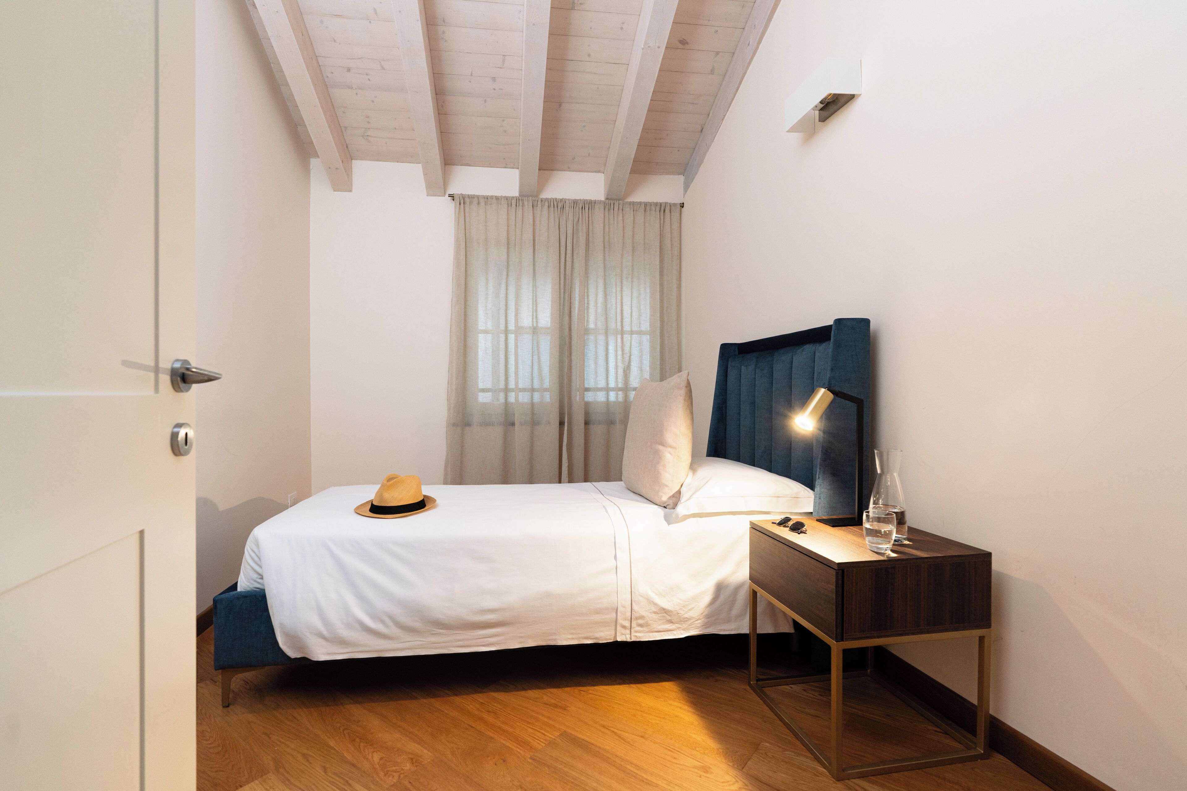 Une chambre avec un plancher en bois, un lit avec une literie blanche, une table de chevet avec une lampe et une fenêtre avec des rideaux.