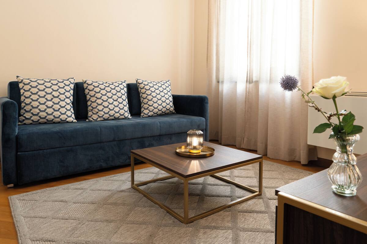 A modern living room with a dark blue sofa, patterned cushions, a glass vase with a white rose, and a geometric coffee table.