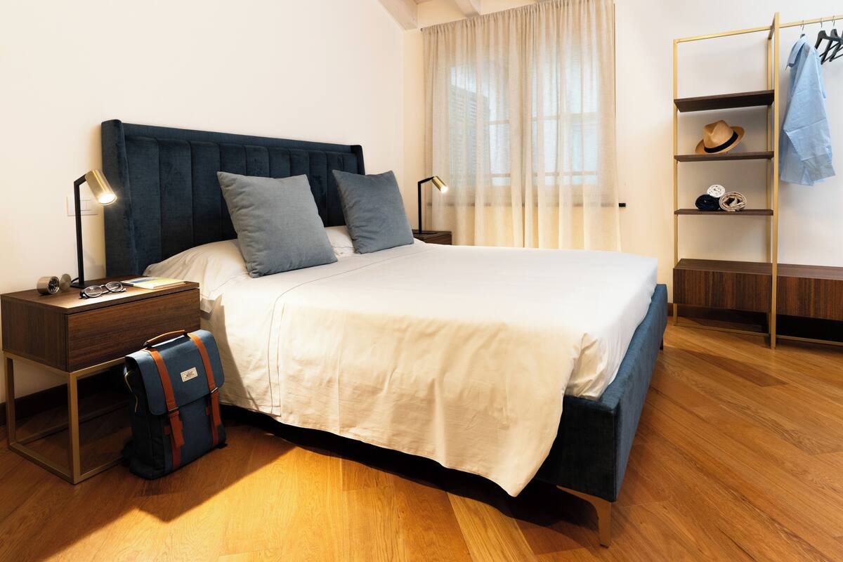 A neatly made bed with a blue headboard, a bedside table with a lamp, a suitcase, and a shelf with a hat and a jacket.