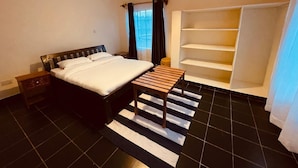 A bedroom with a bed, a nightstand, a wooden bench, a window with curtains, and a built-in closet with shelves.