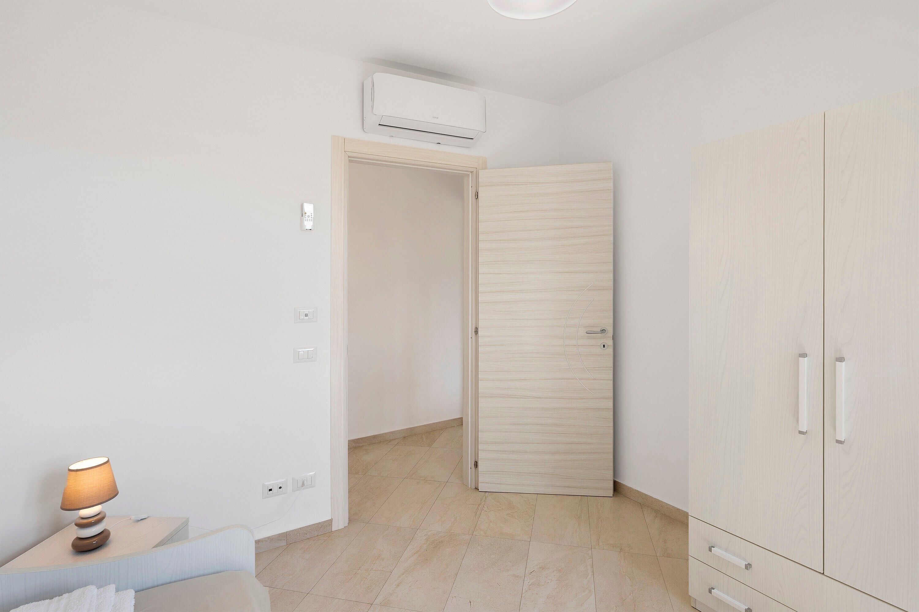 A room with a bed, a bedside table with a lamp, a wardrobe, and an air conditioning unit.