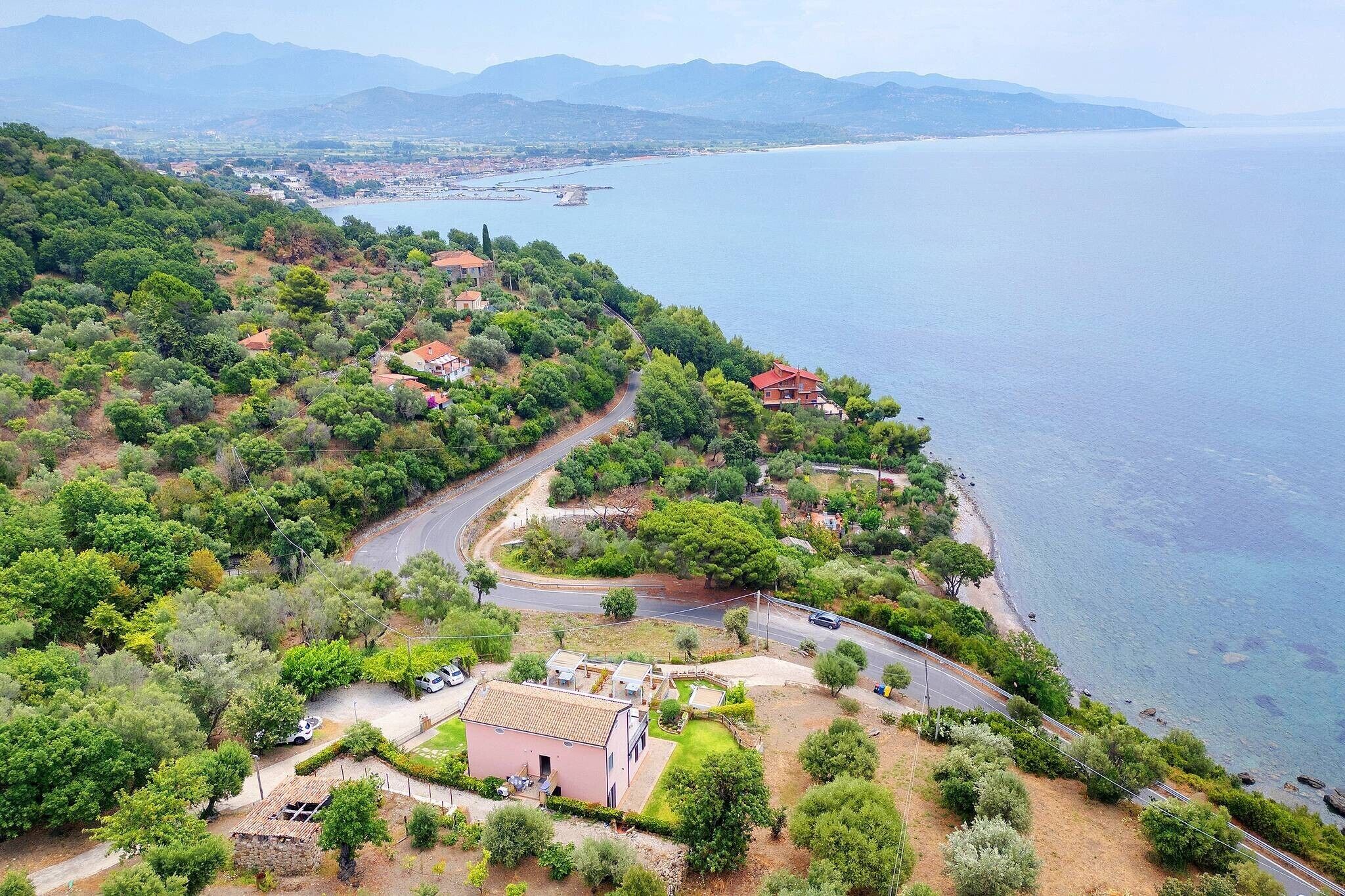 A coastal property with a winding road, a small building, and a view of the sea and distant mountains.