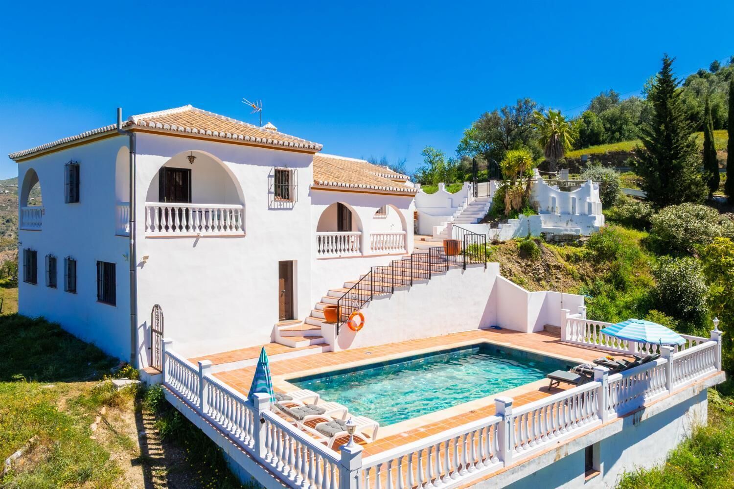 A white villa with a swimming pool, a staircase, and a view of the surrounding greenery.