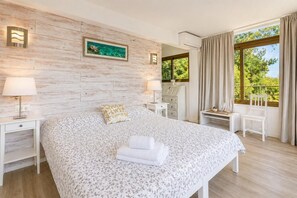A bedroom with a large bed, two bedside tables, a desk, a chair, a window with curtains, and a wall-mounted air conditioning unit.