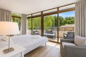 A modern hotel room with a large bed, a bedside table, a lamp, and a view of the balcony with outdoor furniture.