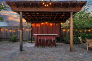 Un espace bar extérieur abrité, avec un mur rouge, des tabourets en bois et des guirlandes lumineuses.