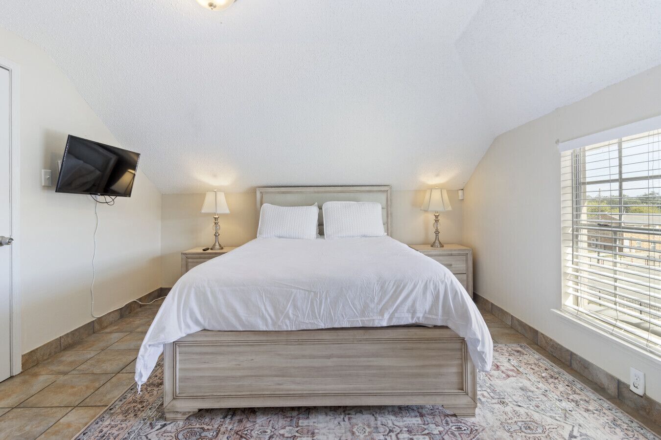 A bedroom with a bed, two bedside tables with lamps, a TV mounted on the wall, and a large window with blinds.