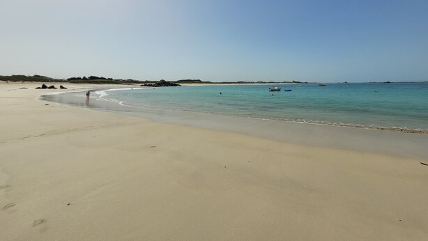 Une plage de sable fin aux eaux calmes, quelques bateaux et, au loin, des formations rocheuses.