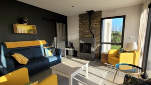 Un salon moderne avec un canapé bleu, des fauteuils jaunes et une table basse. On aperçoit une cheminée au fond, et une grande fenêtre laisse entrer la lumière naturelle.