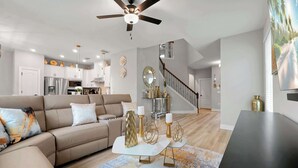 Een moderne woonkamer met een beige bank, een glazen salontafel met gouden details, en een trap die naar een verdieping leidt.