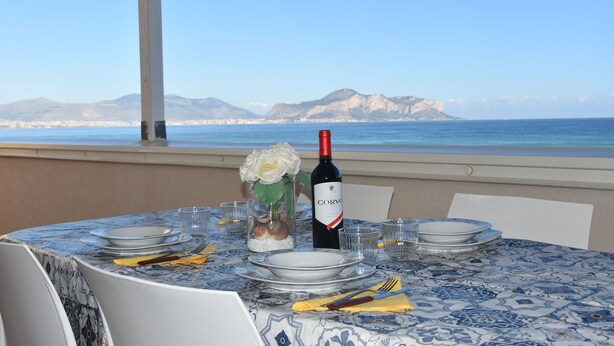 A table set for dining with a bottle of wine, plates, and cutlery, overlooking a coastal view with mountains in the distance. - VILLA HEMINGWAY - ALESSIA'S HOUSE BREATHTAKING SEA VIEW (Ficarazzi)