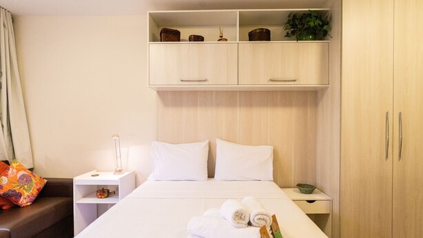 Una habitación de hotel con cama, mesita de noche, estante con objetos decorativos y armario.