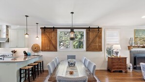 A dining area with a wooden table and chairs, a kitchen with white cabinets, and a large window with wooden shutters. - Hope Cottage sleeps 8 with a heated pool (St Simons Island)