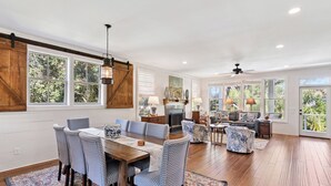 A spacious living area with a dining table, chairs, and a sofa. There are large windows and a ceiling fan. - Hope Cottage sleeps 8 with a heated pool (St Simons Island)