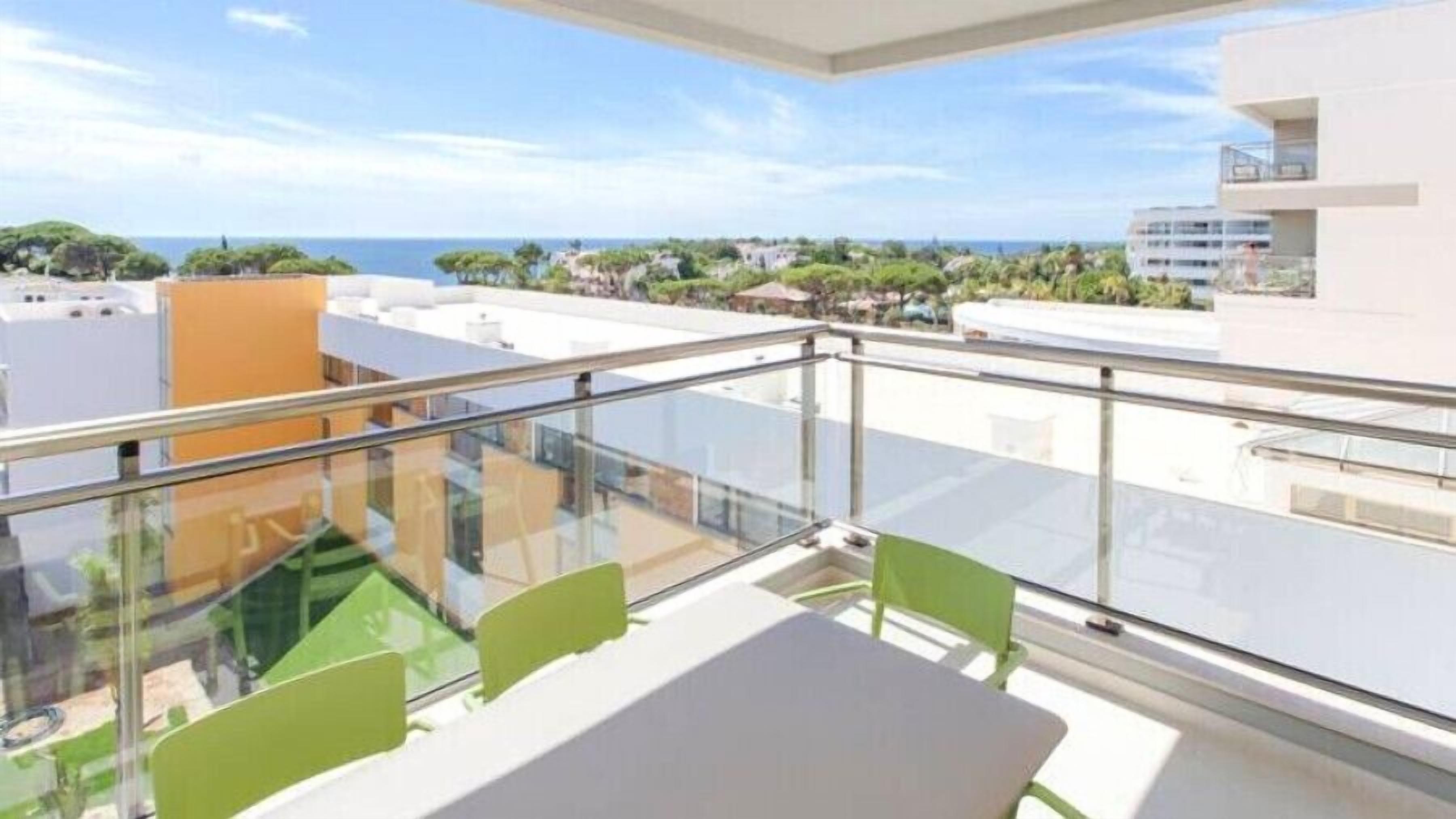 Un balcón con mesa y sillas, con vistas a una zona costera con edificios y vegetación.