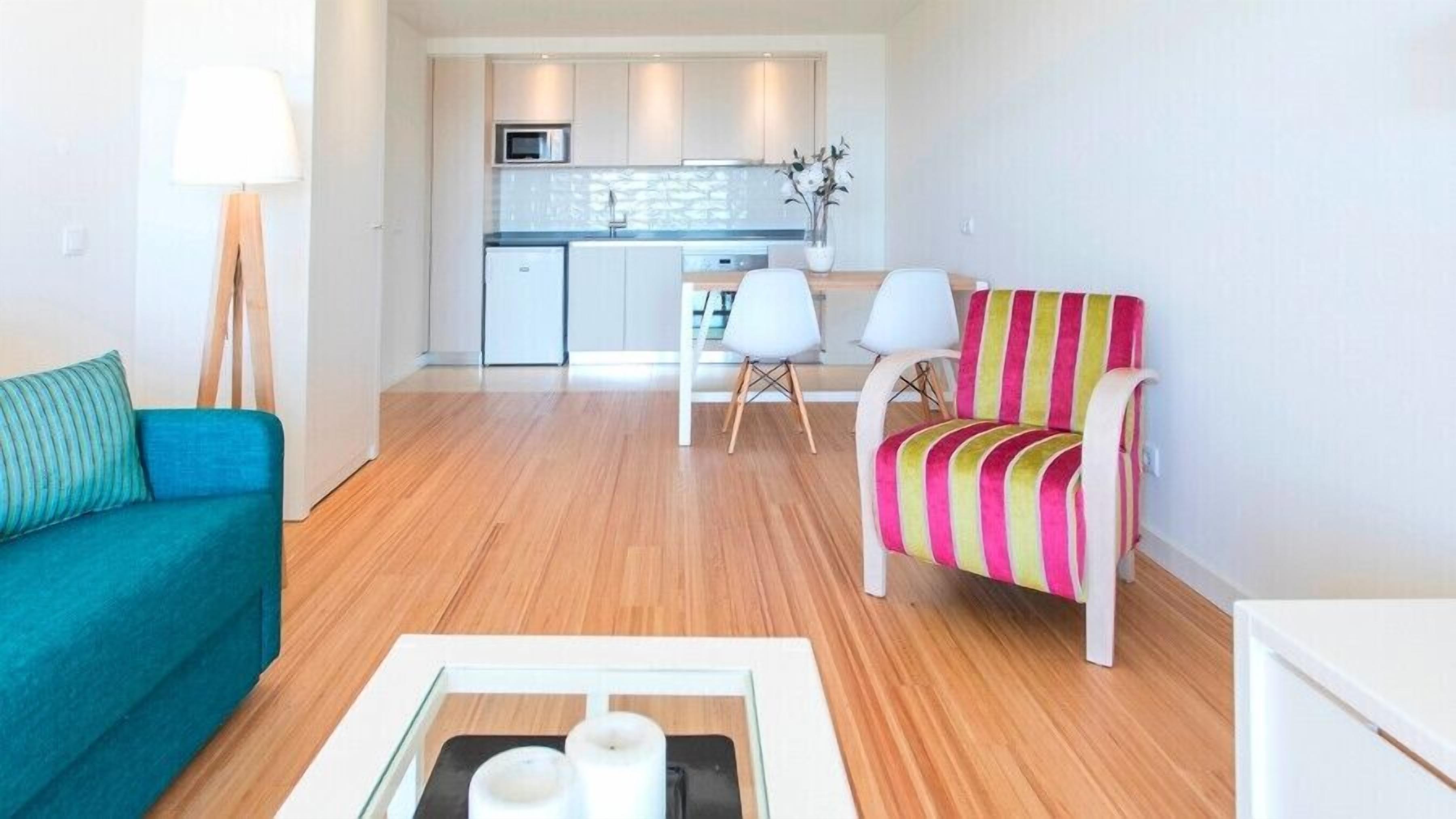 Una sala moderna con un sofá azul verdoso, un sillón colorido a rayas y una mesa de centro de vidrio. Se ve una cocina con armarios blancos y un microondas al fondo.