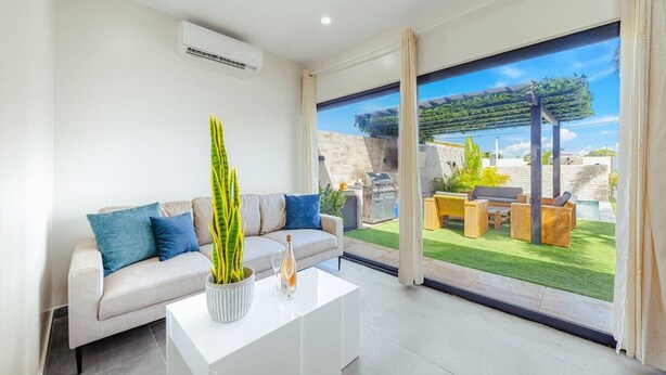 En moderne stue med sofa, sofabord og et stort vindue der giver udsigt til en patio med udendørs møbler og en pool.