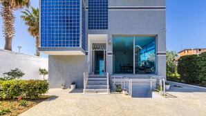 A modern building with a glass entrance, a staircase leading to the entrance, and a paved outdoor area with potted plants. - Apartment With AC and Balcony Golem (Golem)