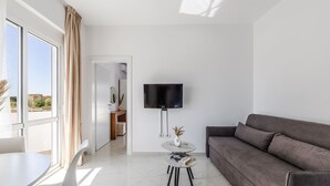 A modern living room with a sofa, a TV, a small table, and a view of the outdoors through large windows. - Apartment With AC and Balcony Golem (Golem)