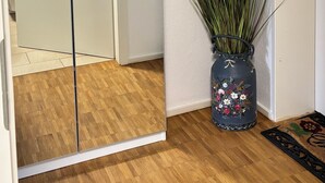 A room with a wooden floor, a mirror reflecting a door, a vase with a floral design, and a wall-mounted light switch.
