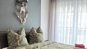 A modern bedroom with a large bed, floral bedding, a decorative wall hanging, and sheer curtains.