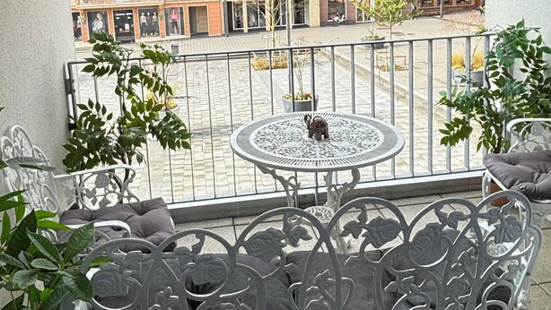 A balcony with a metal table and chairs, potted plants, and a view of colorful buildings.