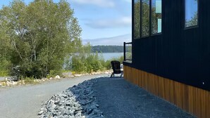 A modern building with large windows overlooking a lake, a chair outside, and a gravel path leading to the entrance. - Glamping Cabin | Best Bay Views | Near Restaurants (Gouldsboro)