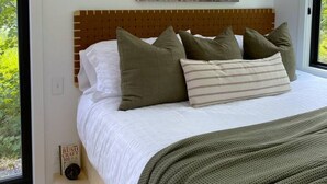 Un dormitorio con cabecera de madera, cama con ropa blanca y varias almohadas, un cuadro en la pared y dos apliques negros.