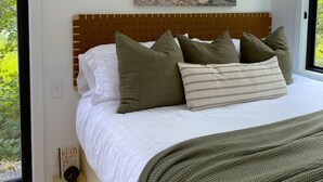 A bedroom with a wooden headboard, a bed with white bedding and multiple pillows, a painting above the bed, and two wall-mounted lamps. - Bay Front A-Frame | Near Birch & Bunker`s Harbor (Gouldsboro)