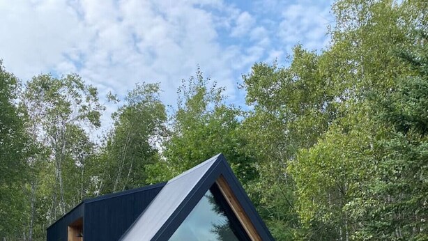 A modern cabin with a triangular roof, wooden exterior, and large windows. It features outdoor seating with Adirondack chairs and a fire pit. - Bay Front A-Frame | Near Birch & Bunker`s Harbor (Gouldsboro)
