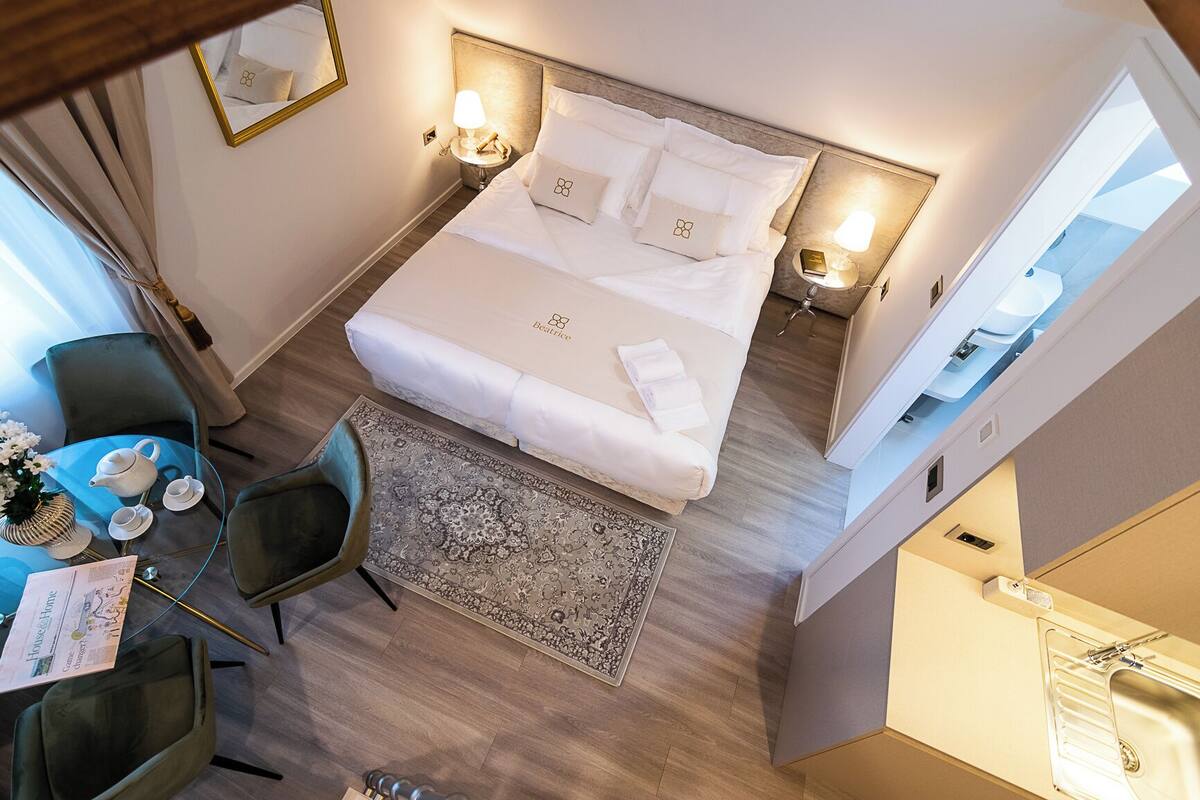Ein Hotelzimmer mit einem Bett, Nachttischen, einem kleinen Tisch mit Teekanne und Tassen, einem Sessel und einem großen Fenster.