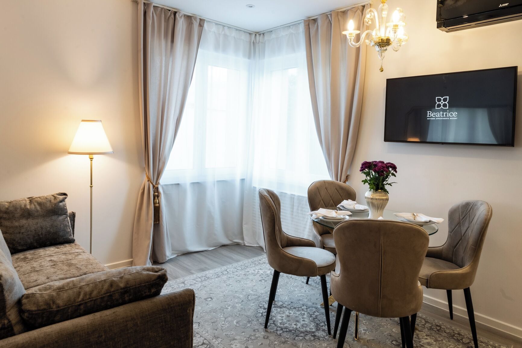 Ein Hotelzimmer mit einer Couch, einem Esstisch mit Stühlen, einer Kristallleuchter, einem Flachbildfernseher und einem Fenster mit Vorhängen.