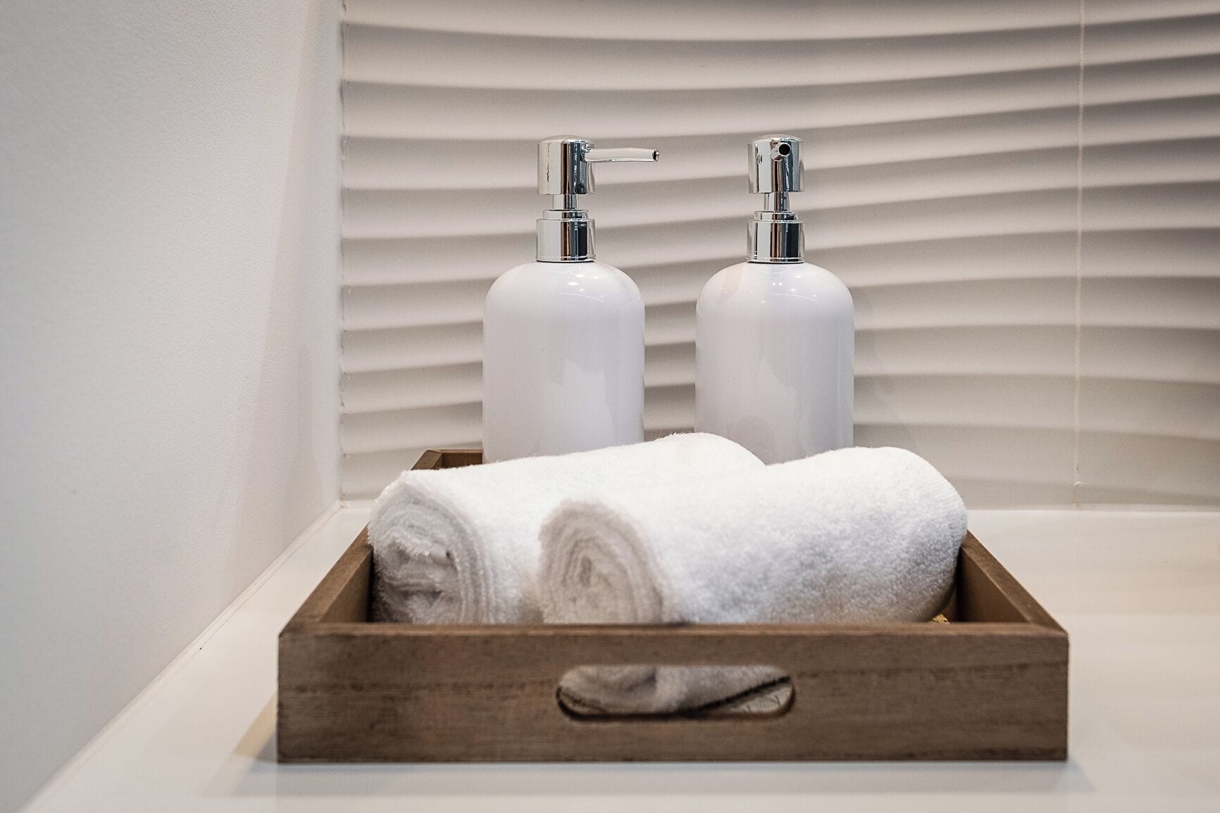 A wooden tray with two white bottles and two rolled towels on a white surface.