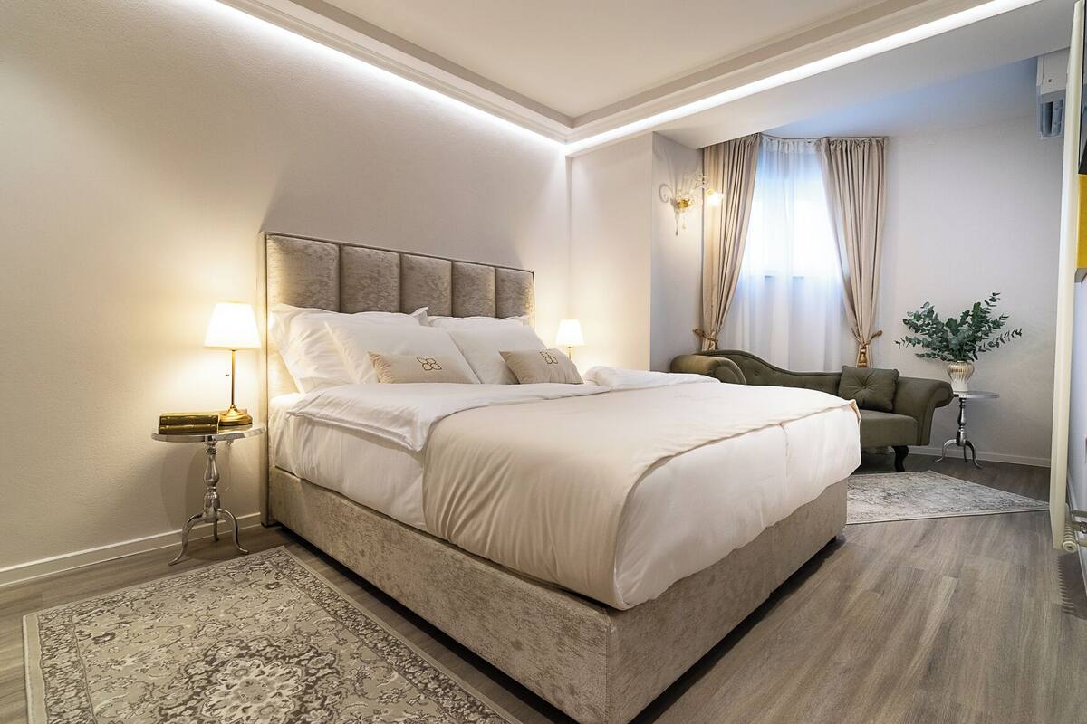 Ein Schlafzimmer mit einem großen Bett, Nachttischlampen, einem Sofa, einem kleinen Tisch mit Pflanze und einem Fenster mit Vorhängen.