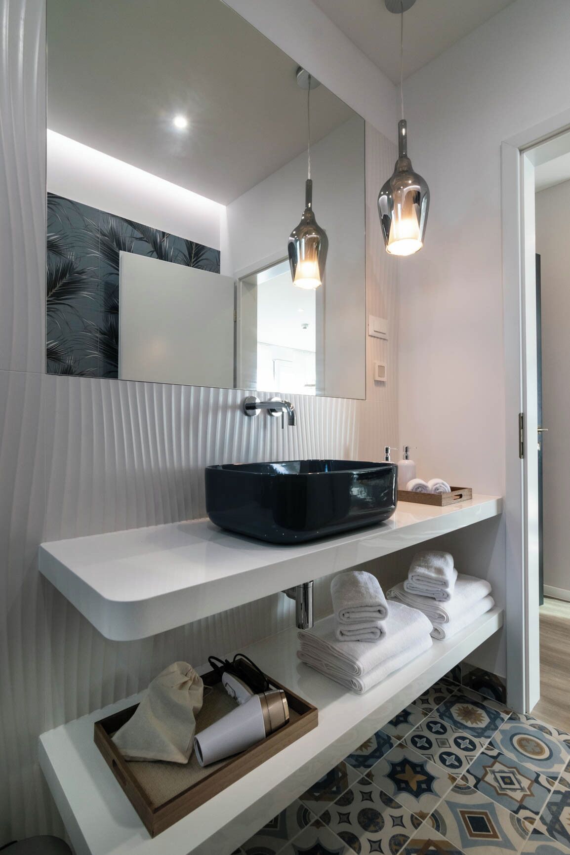 Une salle de bain moderne avec un meuble-lavabo blanc, un lavabo noir et un miroir au-dessus. On y trouve deux lampes suspendues, un porte-serviettes et un carrelage au motif décoratif.
