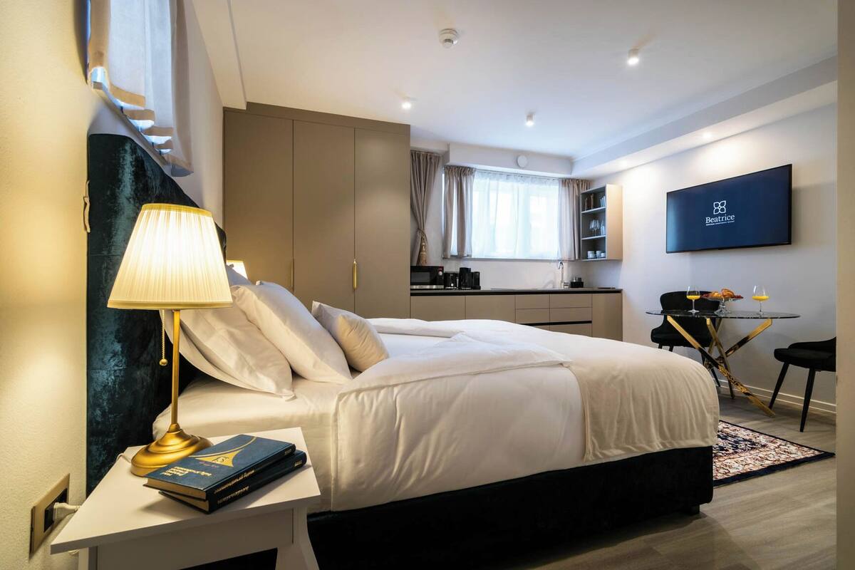 Ein modernes Hotelzimmer mit Bett, Nachttischlampe, Büchern, einem Schreibtisch, einem Fernseher, einer Küchenzeile und einem Esstisch.