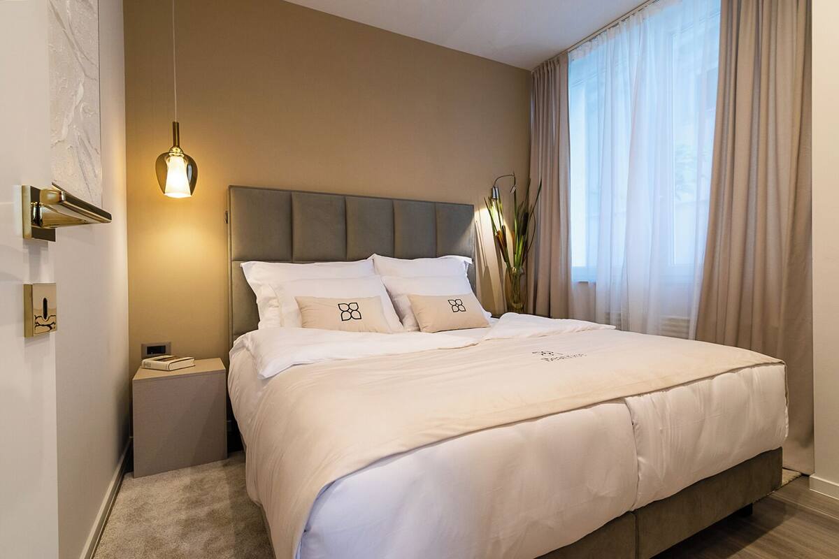 Ein Hotelzimmer mit Bett, Nachttisch, Wandleuchte und einem fensterbepflanzten Fenster.
