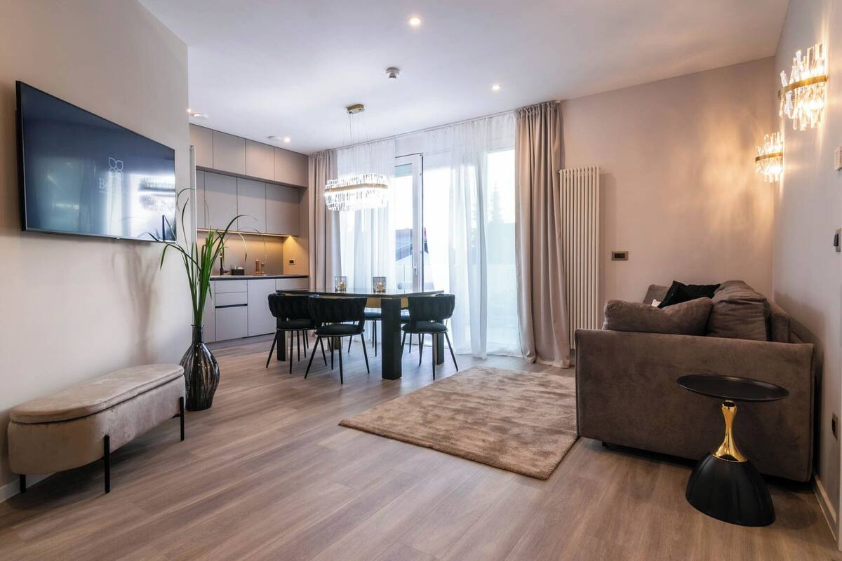 Ein modernes Wohnzimmer mit einer Couch, einem Couchtisch, einem Fernseher, einem Essbereich mit Tisch und Stühlen sowie einer Küche im Hintergrund.