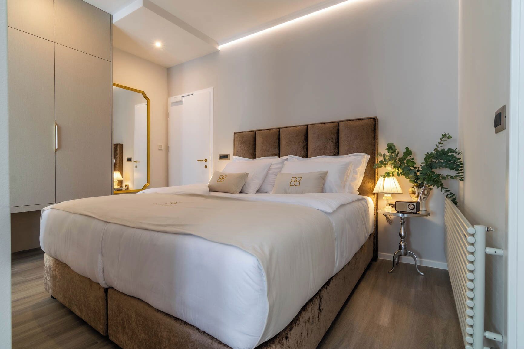A modern bedroom with a large bed, a nightstand with a lamp, a mirror, and a radiator.
