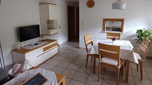 Une pièce comprenant un lit, un téléviseur, une table à manger avec des chaises, une petite table avec un tableau noir et une plante.