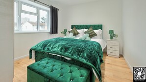 A bedroom with a large bed, a green upholstered bench, a nightstand with a lamp, and a window with curtains.