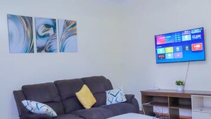 Un soggiorno moderno con un divano grigio, un piccolo tavolino bianco e opere d'arte astratte alle pareti. Un televisore a schermo piatto montato sulla parete mostra diverse piattaforme di streaming.