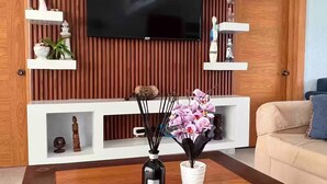 Ein modernes Wohnzimmer mit an einer Holzwand montiertem Fernseher, weißen Regalen mit Dekorationselementen, einem Couchtisch mit Blumenvase und einem beigefarbenen Sofa.