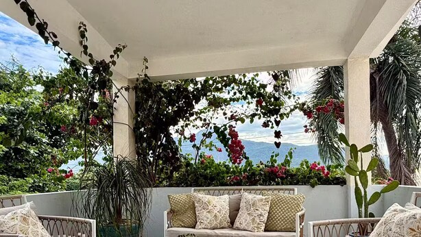 Ein überdachter Außenbereich mit Korbmöbeln, einem runden Glastisch und Blick auf das Meer.