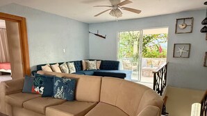 Ein Wohnzimmer mit einem beigefarbenen Ecksofa, einem dunkelblauen Sofa und einem Holztisch. Es gibt einen Deckenventilator, ein Fenster mit Blick auf Grünflächen und eine Tür, die nach draußen führt.
