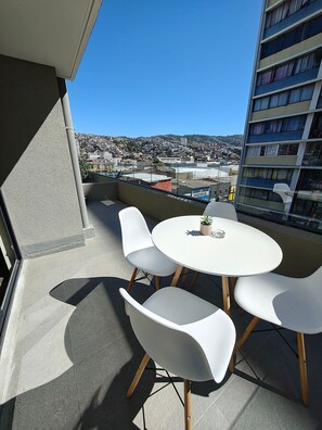 Un balcón con una mesa redonda y cuatro sillas blancas, con vista a un paisaje urbano.